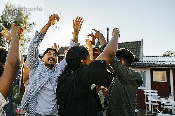 Multiracial group of male and female friends with arms raised dancing in back yard on sunny day at social party