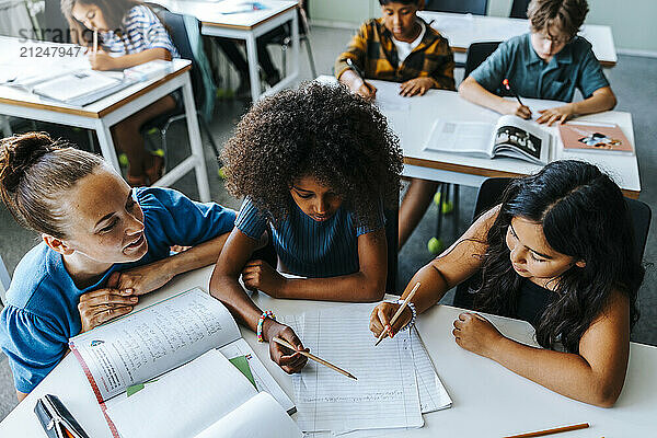 High angle view of teacher helping female pupils studying while sitting near desk in classroom