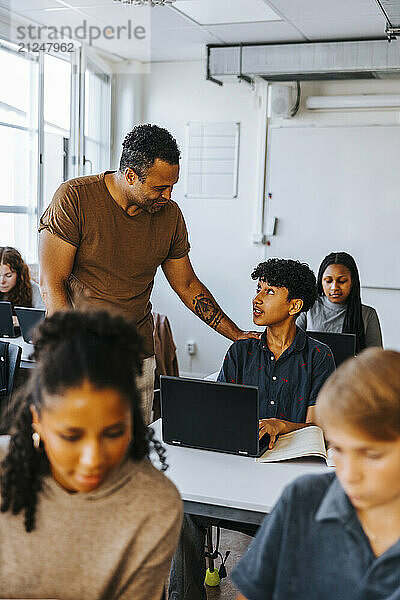 Male professor guiding teenage student siting with laptop in classroom at junior high school