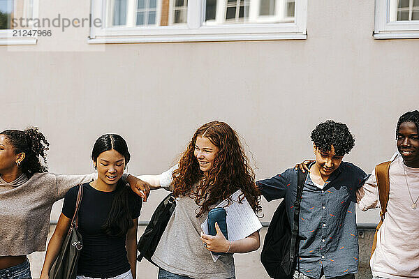 Happy multiracial male and female friends walking with arm around near school building