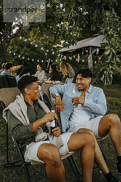 Happy male friend enjoying drinks while sitting on chair and talking in back yard at dinner party