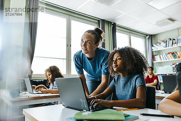 Female teacher bending near curly hair girl sitting with laptop near desk in classroom