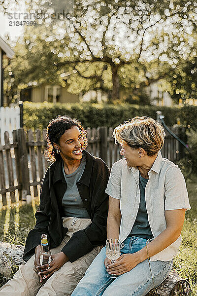 Happy female friends talking with each other while enjoying drinks sitting on log in back yard at sunny day