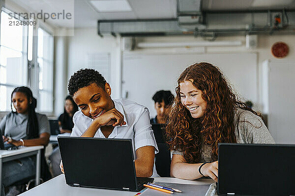 Smiling teenage girl sitting with male classmate while studying on laptop in classroom at school