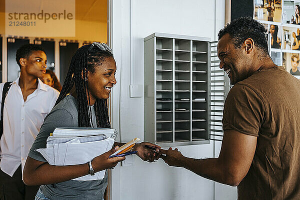 Happy male teacher collecting smart phone from teenage girl standing near doorway of classroom at school