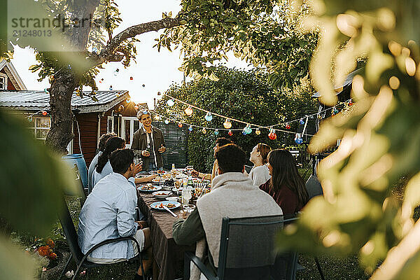 String light decoration with man giving speech to male and female friends at dinner party in back yard