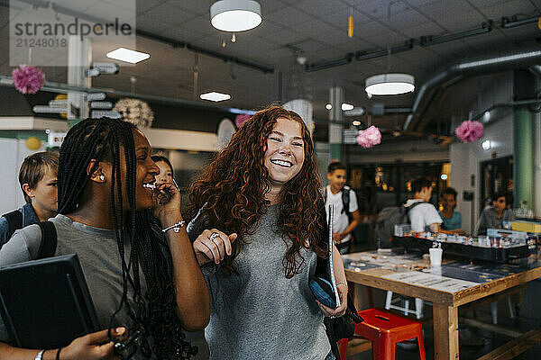 Happy curly haired teenage girl walking with female friend in cafeteria at school