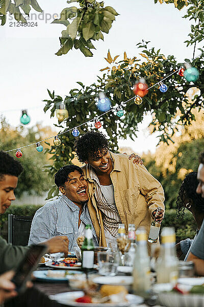 Happy young man embracing male friend sitting near table in back yard at dinner party