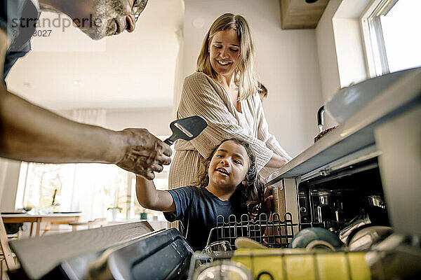 Grandson giving utensil to senior man near dishwasher in kitchen at home