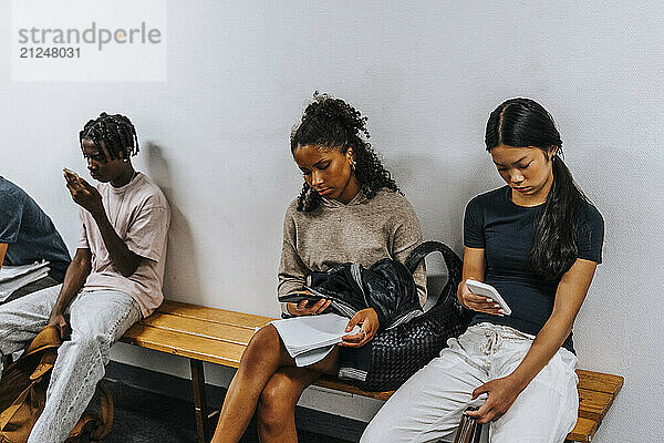 Male and female students using smart phones while sitting on bench near wall at school