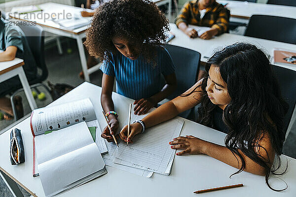 High angle view of female elementary students writing on paper while sitting near desk in classroom at school