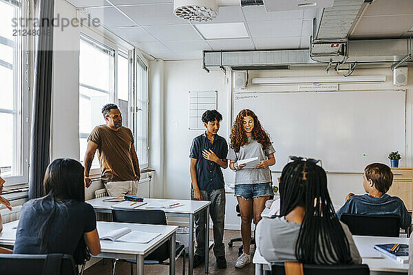 Male professor standing near teenage girl and boy reading in front of junior high students at classroom