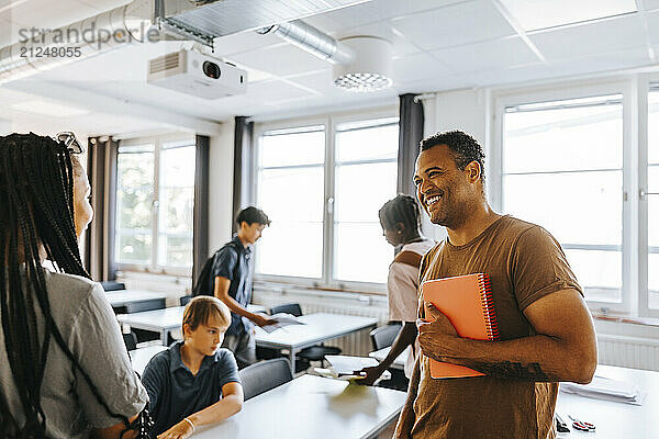 Happy male teacher holding diary while talking with female student in classroom at school