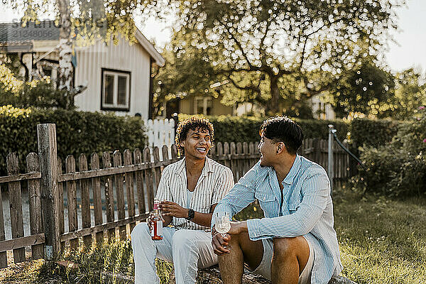 Happy male friends enjoying drinks while sitting in back yard at sunny day