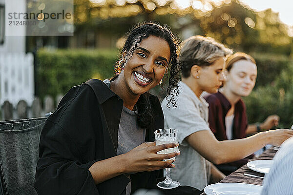 Portrait of happy young woman holding wine glass while sitting near table in back yard for dinner party