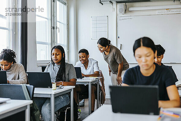 Smiling female teacher assisting student studying on laptop while sitting in classroom at school