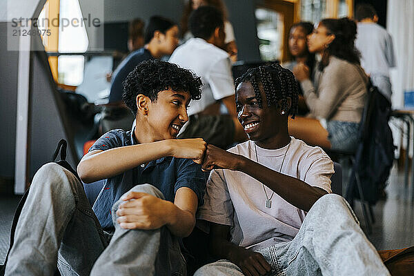 Happy multiracial male friends doing fist bump while sitting in cafeteria at school