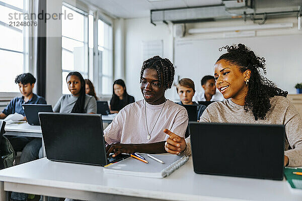 Smiling teenage girl sitting with male classmate while studying on laptop in classroom at junior high school