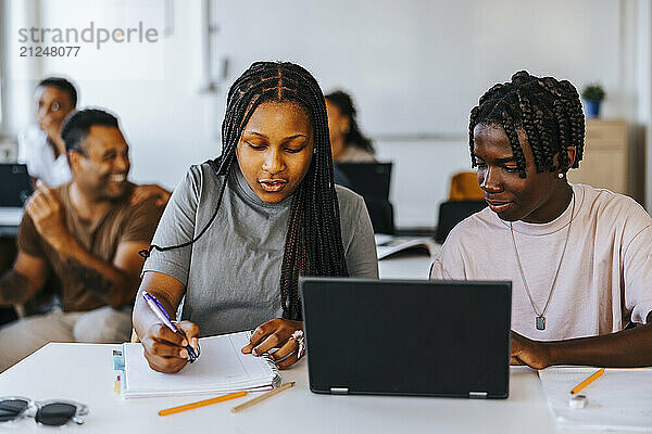 Braided hair girl writing while sitting beside male classmate near desk in classroom at school