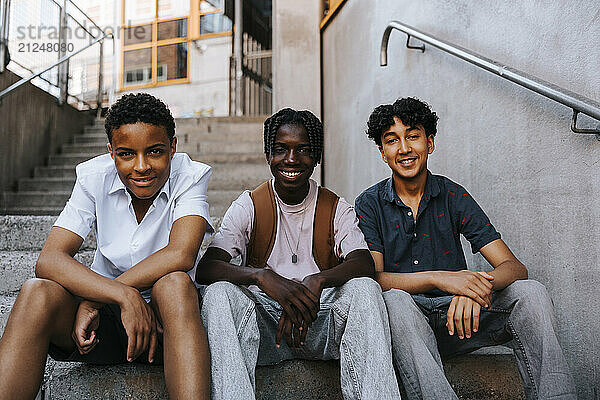 Portrait of happy multiracial male students sitting on stairs of school building