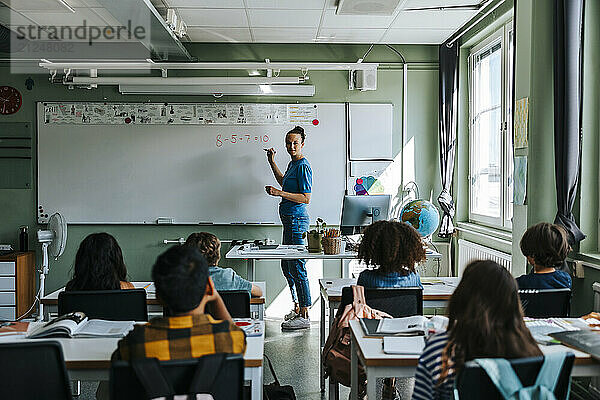 Female teacher teaching mathematics on whiteboard to group of multiracial students in classroom at school