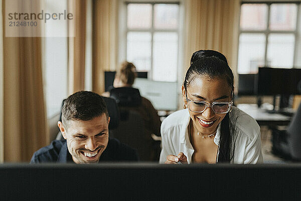 Happy young businesswoman with eyeglasses sharing computer screen with male coworker at workplace