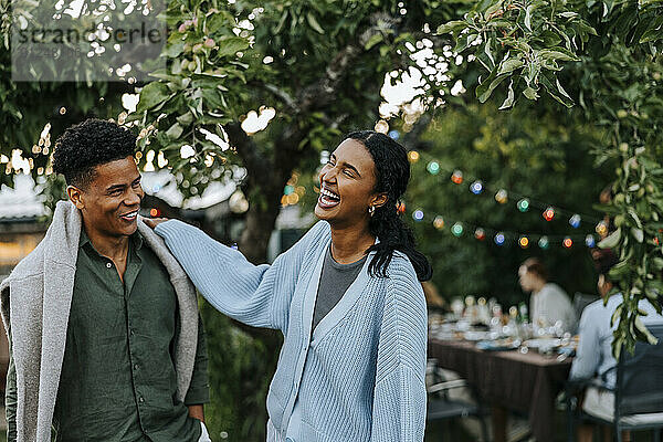 Cheerful young woman with hand on shoulder of male friend standing in back yard at dinner party