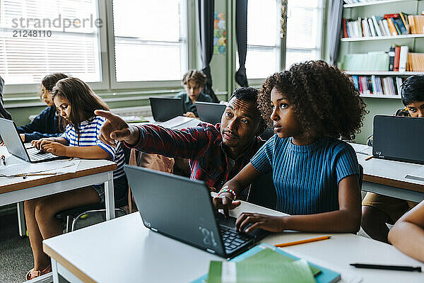 Male teacher pointing while assisting schoolgirl sitting with laptop near desk in classroom