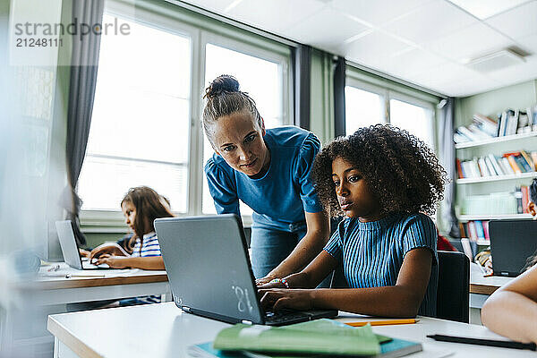 Female teacher assisting curly hair girl studying on laptop while sitting near desk in classroom at elementary school