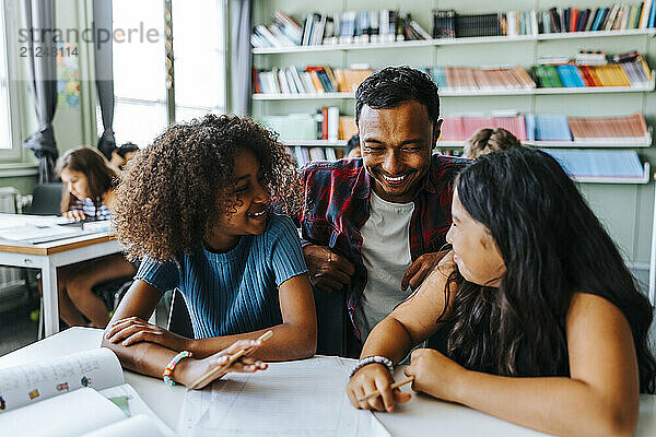 Happy male professor crouching near female pupils sitting near desk in classroom at elementary school