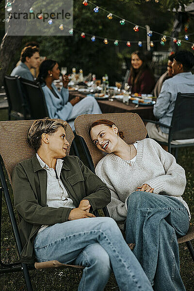 Female friends with legs crossed at knee sitting on chair and talking in back yard at social party