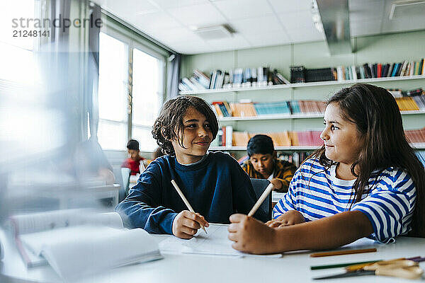 Smiling male and female students holding pencils while sitting at desk in classroom