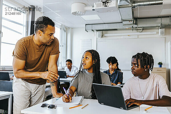 Male professor assisting teenage students sitting with laptop in classroom at junior high school