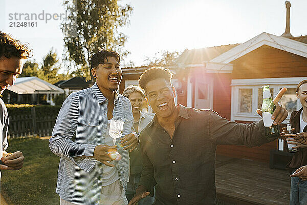 Cheerful male friends enjoying drinks while dancing in back yard on sunny day at party