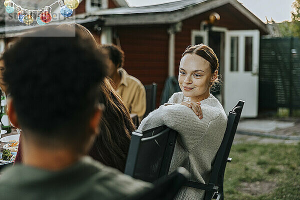 Smiling young woman leaning on chair while talking with male friend in back yard at dinner party