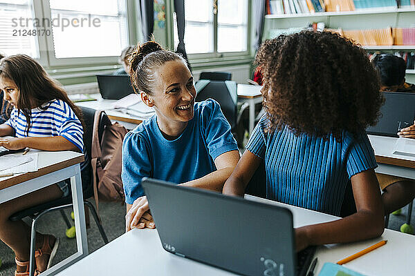 Happy female professor beside curly hair student doing e-learning in classroom at elementary school