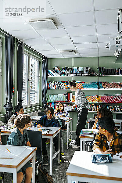 Side view of female professor giving test papers to elementary students sitting in classroom at school