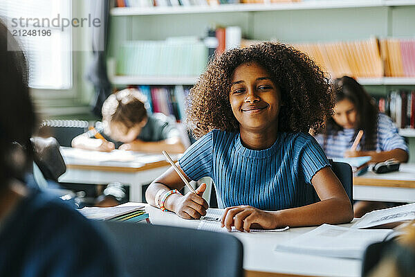 Portrait of smiling curly hair student holding pencil while sitting near desk in classroom at school