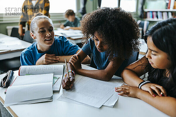 Professor crouching near female student writing on paper while sitting beside classmate in classroom at elementary schoo