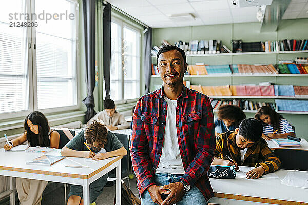 Portrait of smiling male teacher in plaid shirt sitting on desk in classroom at school