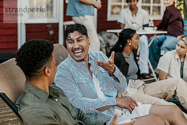 Cheerful young man enjoying drinks with male friend while sitting on chair in back yard at social party