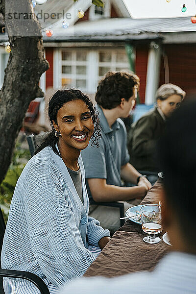 Smiling young woman talking with friend while sitting near dining table in back yard at social party