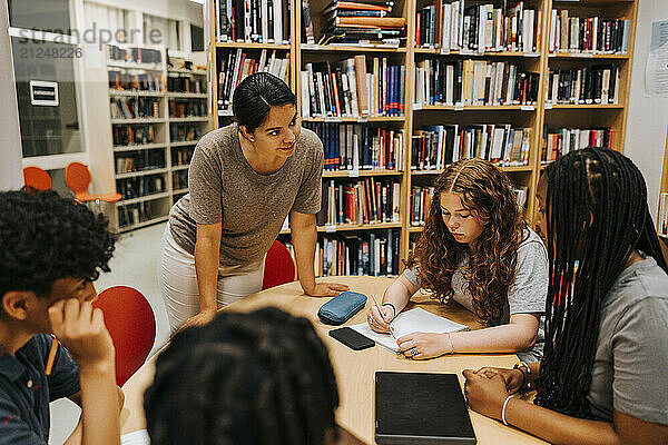 Female librarian assisting teenage students sitting near table in library at school