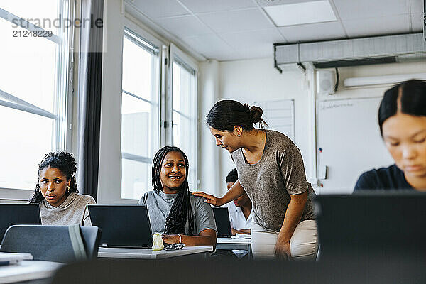 Female teacher assisting student sitting with laptop in classroom at junior high school