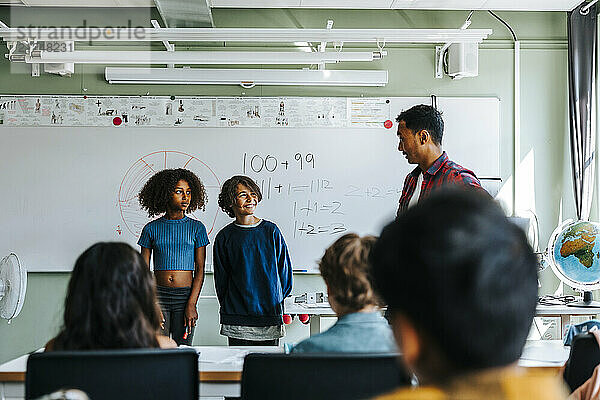 Teacher talking with male and female students standing near whiteboard in classroom