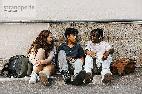 Male and female teenage friends discussing while sitting near wall of school building