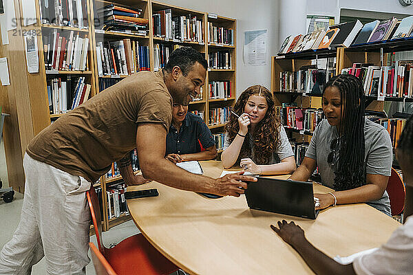 Happy male librarian assisting teenage students sitting near table in library at school