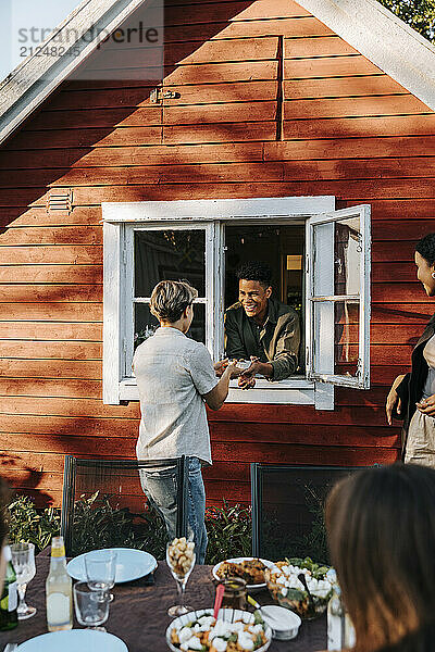 Happy young man passing food through window to female friend standing in back yard at dinner party