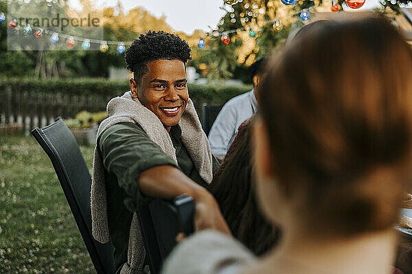 Smiling young man sitting on chair while talking with female friend in back yard at dinner party