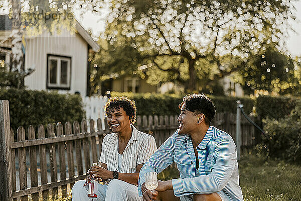 Happy male friends talking while enjoying drinks sitting in back yard at sunny day